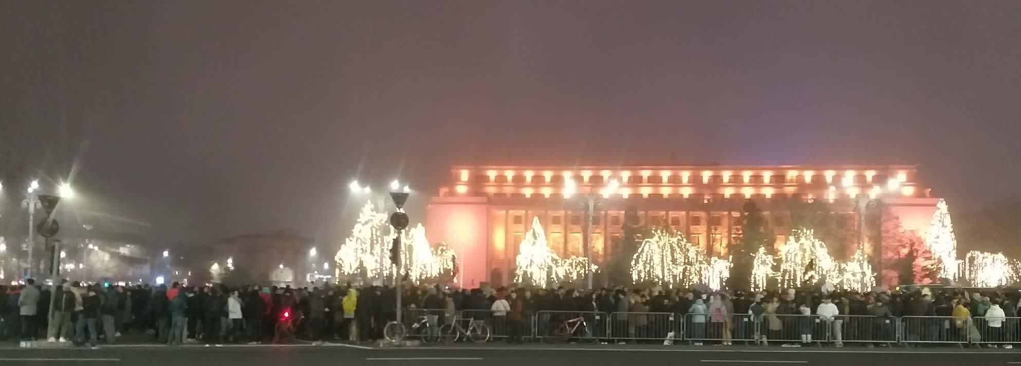 Mulțime densă de protestatari adunați în Piața Victoriei noaptea, în fața clădirii Guvernului iluminată în roșu, cu decoruri luminoase sărbătorești și bariere de siguranță.